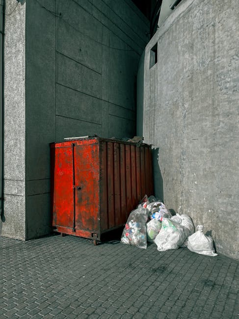 An outdoor scene showing a large, red, metal waste skip with visible signs of rust and wear, positioned against a grey concrete wall next to a dark green wall with horizontal paneling. Several tied plastic rubbish bags, predominantly white with some transparent sections revealing contents such as paper and plastic bottles, are stacked beside the skip on the ground, which is paved with textured grey bricks. The setting appears to be an urban alleyway or service area, where independent waste collection or on-site rubbish clearance is being carried out by Rubbish Removal Finchley, illustrating a typical scene of bulk waste disposal outside a commercial or residential building.