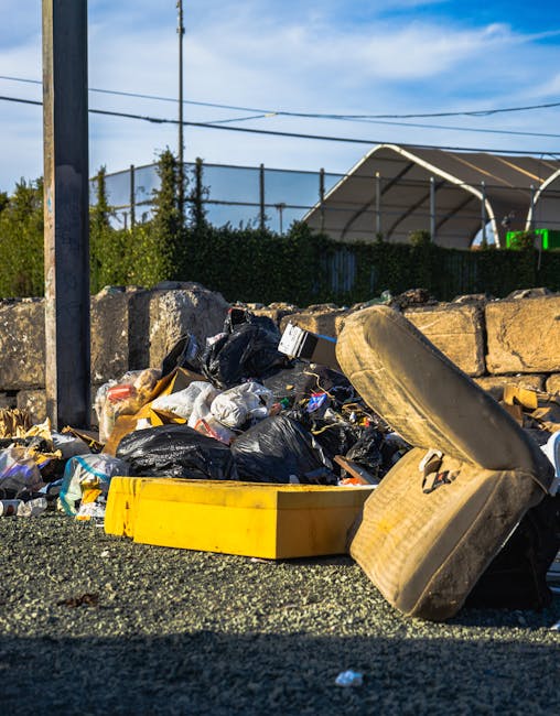 An outdoor scene showing a large, red, metal waste skip with visible signs of rust and wear, positioned against a grey concrete wall next to a dark green wall with horizontal paneling. Several tied plastic rubbish bags, predominantly white with some transparent sections revealing contents such as paper and plastic bottles, are stacked beside the skip on the ground, which is paved with textured grey bricks. The setting appears to be an urban alleyway or service area, where independent waste collection or on-site rubbish clearance is being carried out by Rubbish Removal Finchley, illustrating a typical scene of bulk waste disposal outside a commercial or residential building.