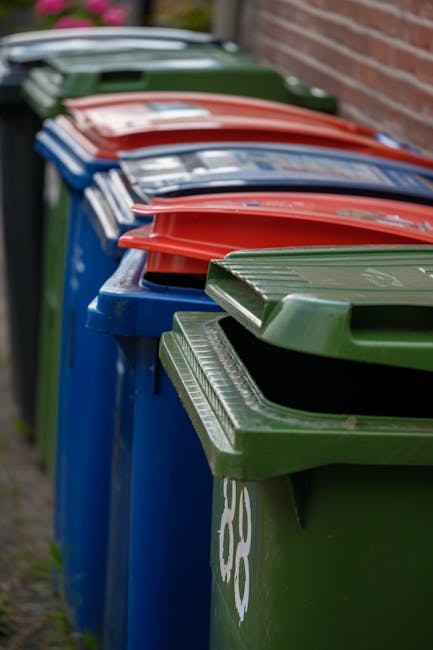 A row of waste bins placed along a pavement outside a brick building, consisting of green, blue, red, and dark green containers. The green bin in the foreground has a textured lid with ridges and is marked with a white recycling symbol. The blue bins behind it have smooth lids, while the red bins have matching smooth lids, and the dark green bin at the back features a similar textured lid. The bins are aligned closely together, with some lids partially overlapping. The environment appears to be an outdoor urban or residential area with natural daylight illuminating the scene, highlighting the colors and textures of the various bin materials. The image emphasizes the importance of suitable waste storage solutions for household or commercial rubbish management, consistent with waste collection practices described by Rubbish Removal Finchley and related to alternative methods such as private disposal or on-site clearance, where bins are positioned for easy access before scheduled collection.