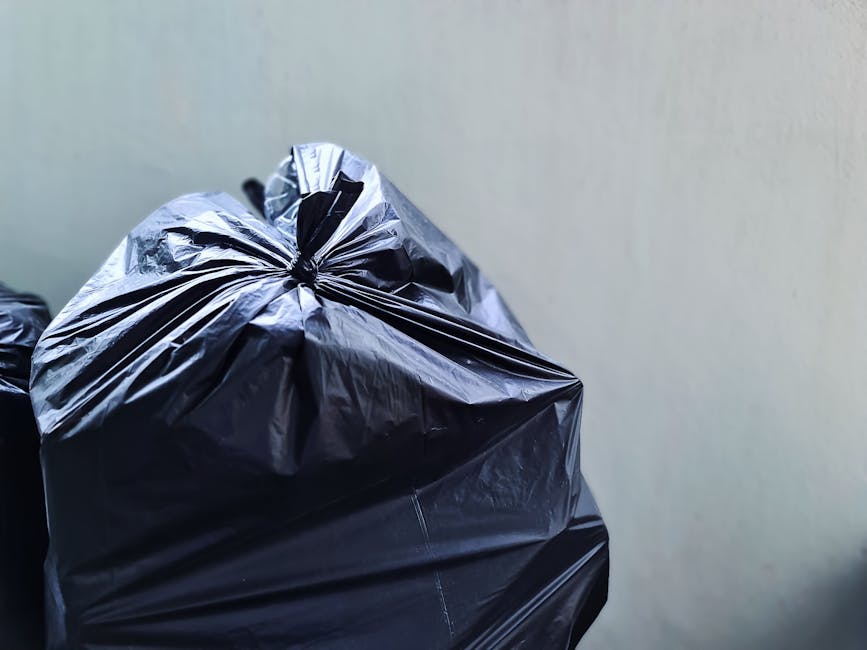 Close-up view of three black garbage bags made of shiny plastic material, tightly knotted at the top, placed against a plain, light-colored wall. The bags are filled with waste and sit on an outdoor surface, possibly a driveway or pavement, with another partially visible bag in the background. The scene reflects a typical private waste collection or rubbish removal activity, with the bags representing bulk or excess rubbish awaiting collection by a service like Rubbish Removal Finchley. The lighting is natural, highlighting the glossy texture of the bags and emphasizing their fullness. The environmental context suggests an on-site waste handling scenario, aligned with non-council-based rubbish disposal options.