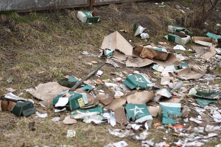 A patch of outdoor ground covered with various discarded waste materials, including flattened cardboard boxes, some with green and white printed logos, crumpled and torn, scattered across a grassy and dirt area. Some cardboard pieces are leaning against a raised wooden retaining wall at the top of the image, which is partially visible. Among the debris, there are small bits of paper and plastic waste, as well as a broken plastic pipe and some unidentifiable small objects. The environment appears unkempt, with waste strewn along the ground and surrounding area, illustrating typical scenes where independent waste disposal might be needed. This image aligns with the context of rubbish removal services, such as those offered by Rubbish Removal Finchley, which deal with clearing waste from outdoor spaces. Natural lighting suggests an overcast day, with no visible shadows, creating a neutral and factual atmosphere suitable for informative or service-related content about rubbish clearance options in the North Finchley area.