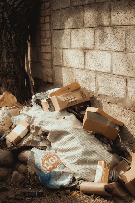 A pile of discarded cardboard boxes, some flattened and others partially opened, rest against a rough brick wall outdoors near a tree trunk. The boxes display various print and branding in shades of brown, beige, and black, with some torn or crumpled. Among the boxes is a large, loosely wrapped white sack that appears to contain additional waste materials, and a blue plastic bag with orange lettering. The ground is composed of dirt and small stones, with uneven terrain and scattered debris. The scene captures an area suitable for private waste collection or on-site rubbish removal services, with the cardboard and refuse organized for disposal. The background brick wall shows signs of weathering, and the natural light indicates daytime, highlighting the textures and colors of the waste materials. This setup illustrates typical conditions where independent rubbish collection might be employed for clearing clutter from outdoor spaces near residential or commercial buildings, supported by companies such as Rubbish Removal Finchley.