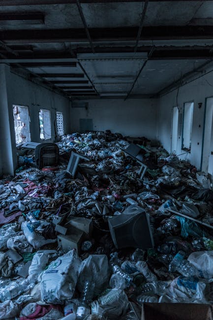 The image depicts an indoor space, likely a room or hall, filled with an extensive accumulation of mixed rubbish and discarded items covering the entire floor. The waste includes numerous crumpled plastic water bottles, crumpled paper, and scraps of packaging, reflecting a variety of materials with different textures such as transparent or opaque plastics and paper. Several old computer monitors and electronic equipment are scattered among the debris, some leaning or toppled over, with casings and screens visibly damaged or cluttered. The room has plain, light-colored walls with windows along the left side allowing natural light to slightly illuminate the scene, revealing a ceiling with exposed beams and some ceiling panels missing. Overall, the environment appears unmanaged, with no clear arrangement or disposal method evident, indicating an urgent need for professional rubbish removal services from companies like Rubbish Removal Finchley to manage on-site clearance or alternative waste handling solutions in such cases.