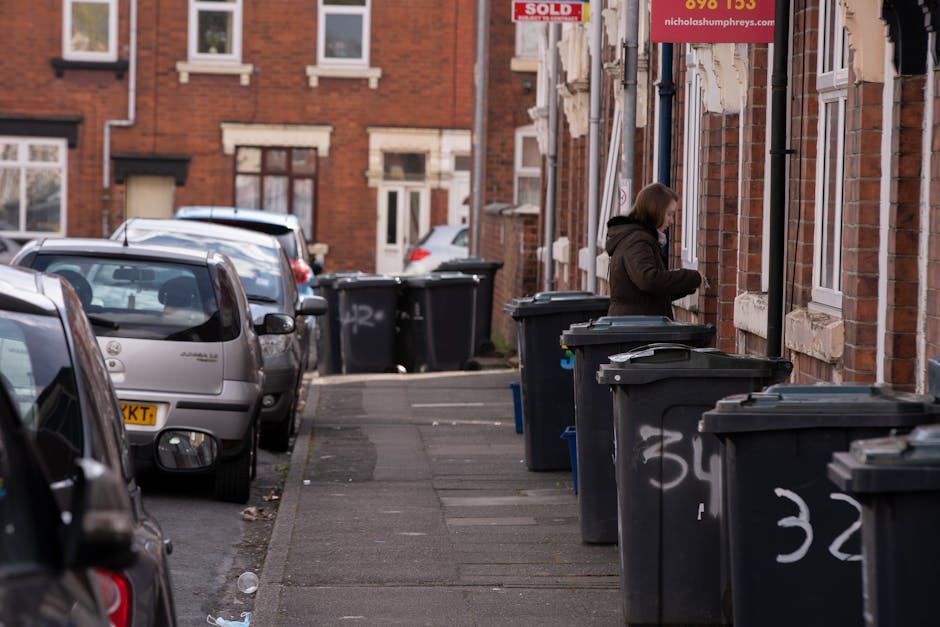 A row of black wheelie bins with closed lids are aligned along the edge of a residential pavement on a city street. Each bin features white painted numbers, with the number 32 visible in the foreground. The bins are positioned right next to a brick building with multiple white-framed windows that have ledges beneath them. A woman dressed in a dark hooded coat stands beside one of the bins, appearing to be in the process of managing rubbish disposal or collection. The street is lined with parked cars on the left, including a silver hatchback closest to the camera, extending down the street where additional vehicles are visible, with some partially obscured by the bins. In the background, the street extends further, featuring more brick houses and parked cars. Environmental lighting suggests an overcast day, typical of urban waste management settings, with a realistic depiction of private waste disposal arrangements facilitated by rubbish removal services such as Rubbish Removal Finchley.