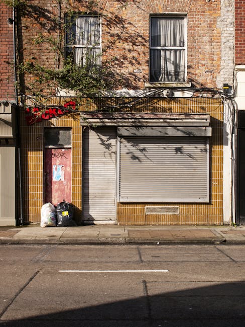 The image depicts the front exterior of a two-storey brick building with a ground-floor commercial unit that appears to be vacant or closed, featuring a closed metal roller shutter door and a smaller metal door to its left, both painted in a light grey tone with visible wear. To the left of the doors, there are three black and white garbage bags and a white plastic bag placed on the pavement, indicating rubbish awaiting collection. The façade around the entrance has yellowish-brown ceramic tiles, with some graffiti on the lower red-painted wall section adjacent to the bags. Above, the upper floor has two sash windows with closed curtains, framed by red brickwork and partially shaded by the branches of a tree, which cast shadows across the building's surface. Electrical wires are mounted on the wall, running horizontally and a small air conditioning unit is visible beneath the upper windows. The scene suggests a typical urban building in need of waste removal or clearance services, aligning with private rubbish collection practices often employed by local businesses or property owners such as Rubbish Removal Finchley, servicing the Finchley area on High Road.
